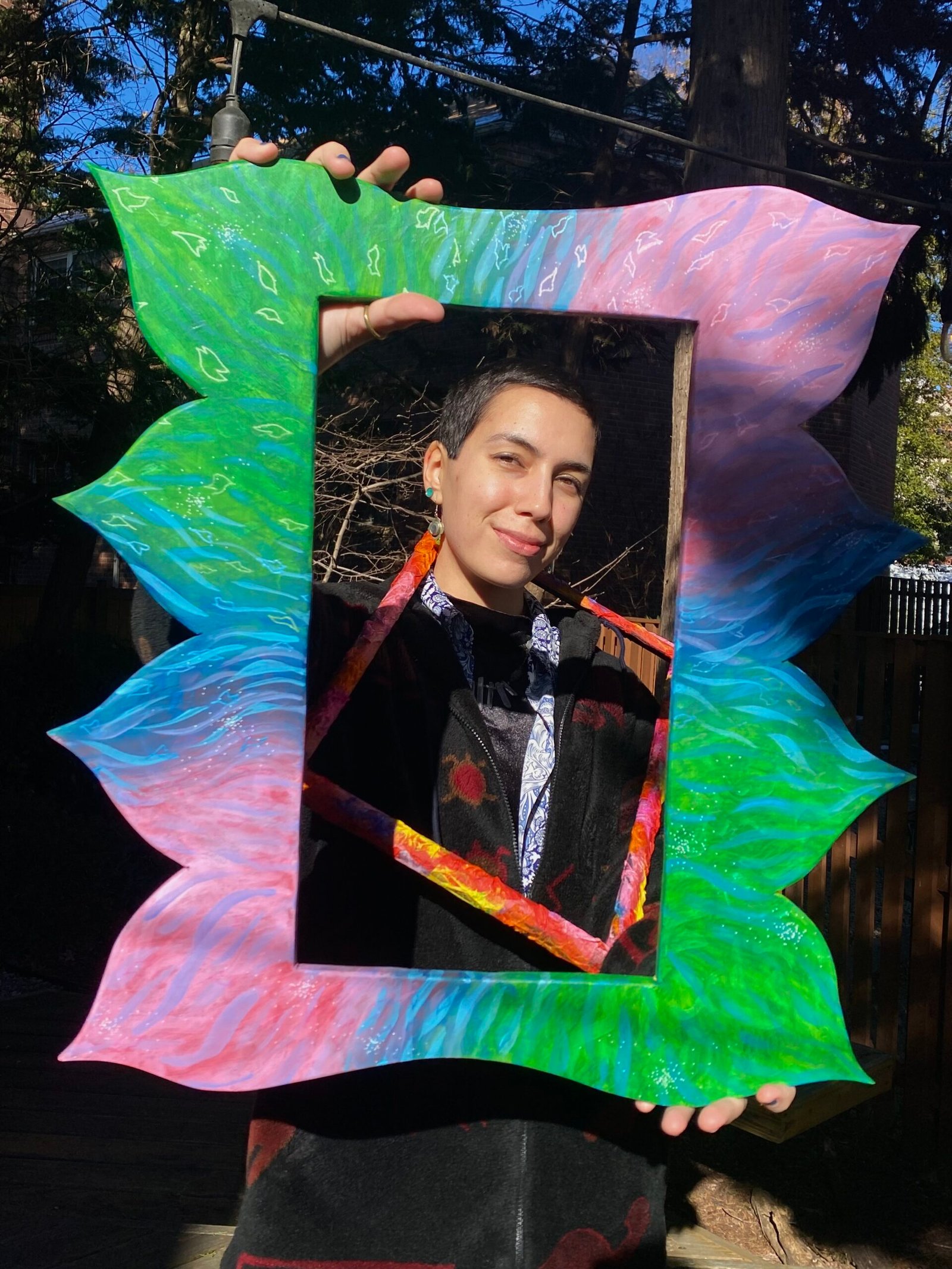 A non-binary latine person with a shaved head holds up a frame and smiles behind it.