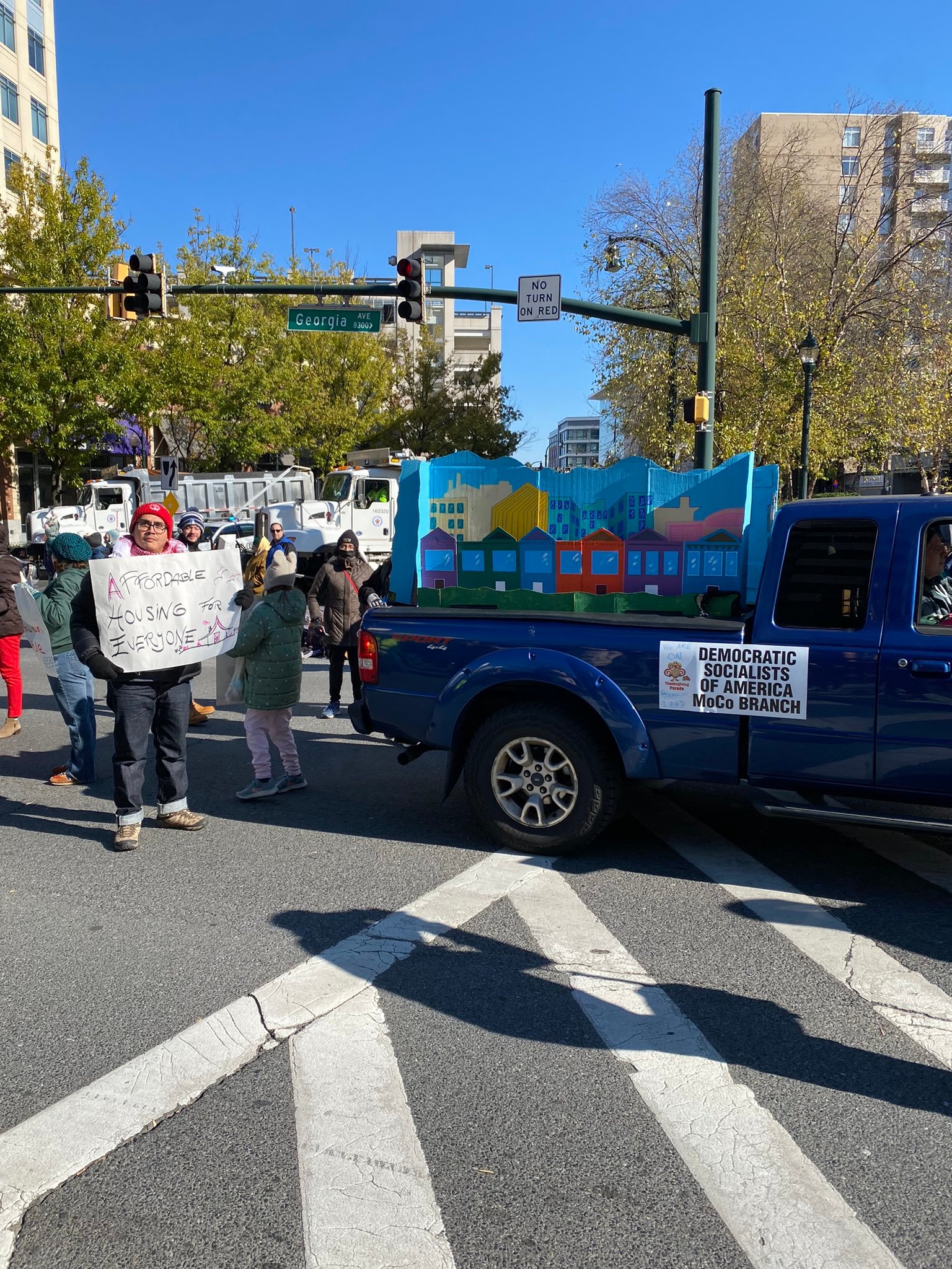 Rent stabilization parade group hold signs and stand next to a blue truck with a float of a cardboard city.