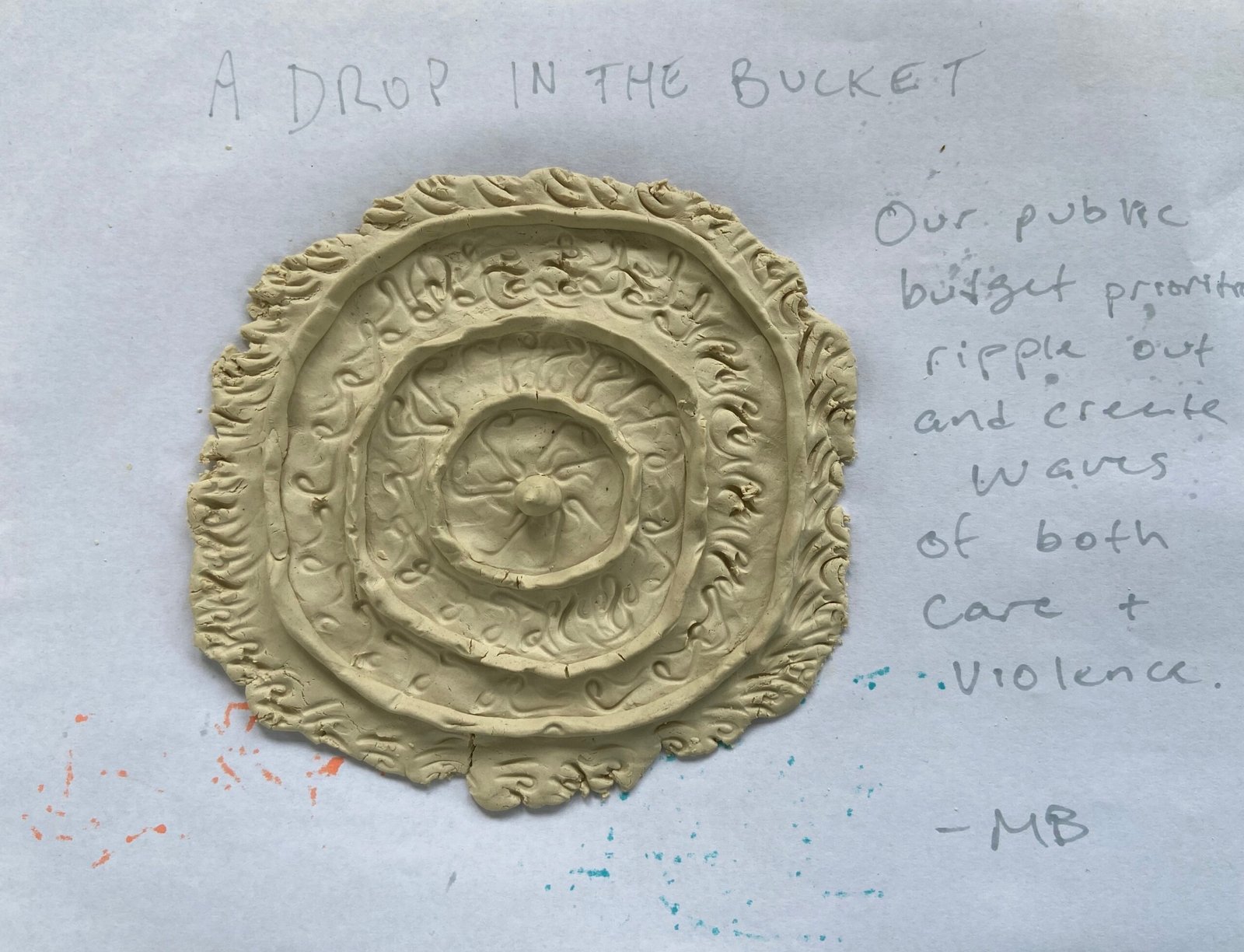A piece of paper with a clay representation of ripples on water. The handwriting reads a drop in the bucket. Our public budget prioritizes a ripple out and creates waves of both care and violence.